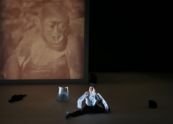 A baby monkey is on a projection screen behind the stage as a actress is doing the splits in front of it.