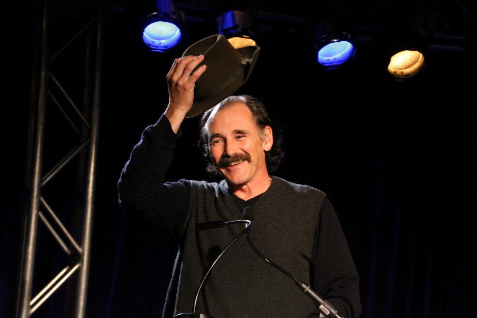 A man takes his hat off on stage in front of a microphone. Blue and yellow spot lights are above him