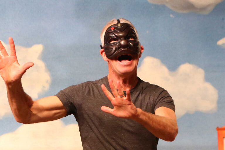 A man with a black mask stands in front of a blue sky with white clouds. HIs mouth is open and his hands up open