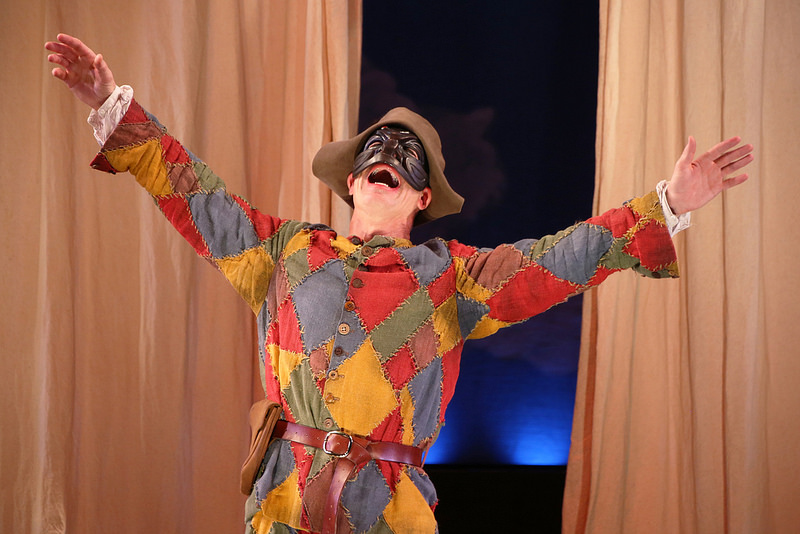 A scarecrow standing in front of half closed curtains with his hands raised above his head as he laughs