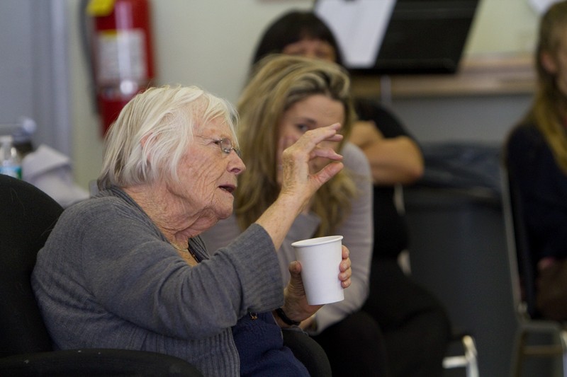A older lady sits with a cup in one hand and her other hand is held up as she pinches her pointer finger and thumb together