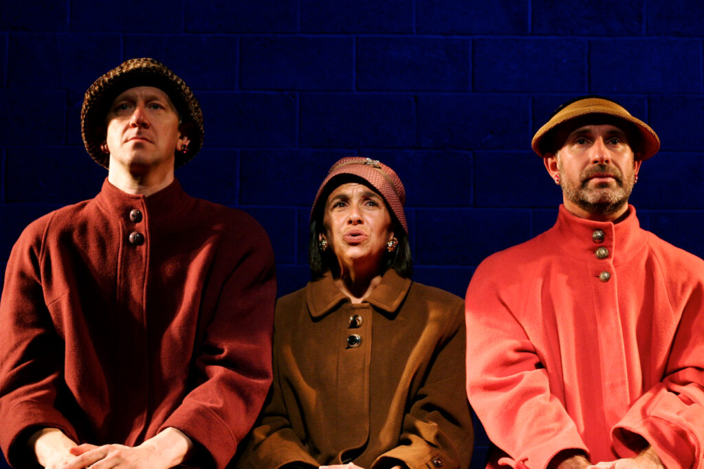 Three actors dressed in warm coats and all wearing hats sit with a blank look on their face