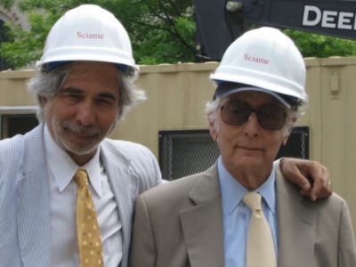Two men stand with white hard hats on