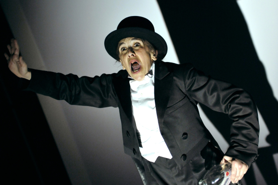A Actress dressed in a black tux and black top hat stands with her mouth open and one hand up in the air