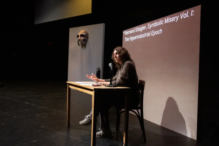 A woman sitting at a desk with a microphone answering questions with a projection screen behind her.