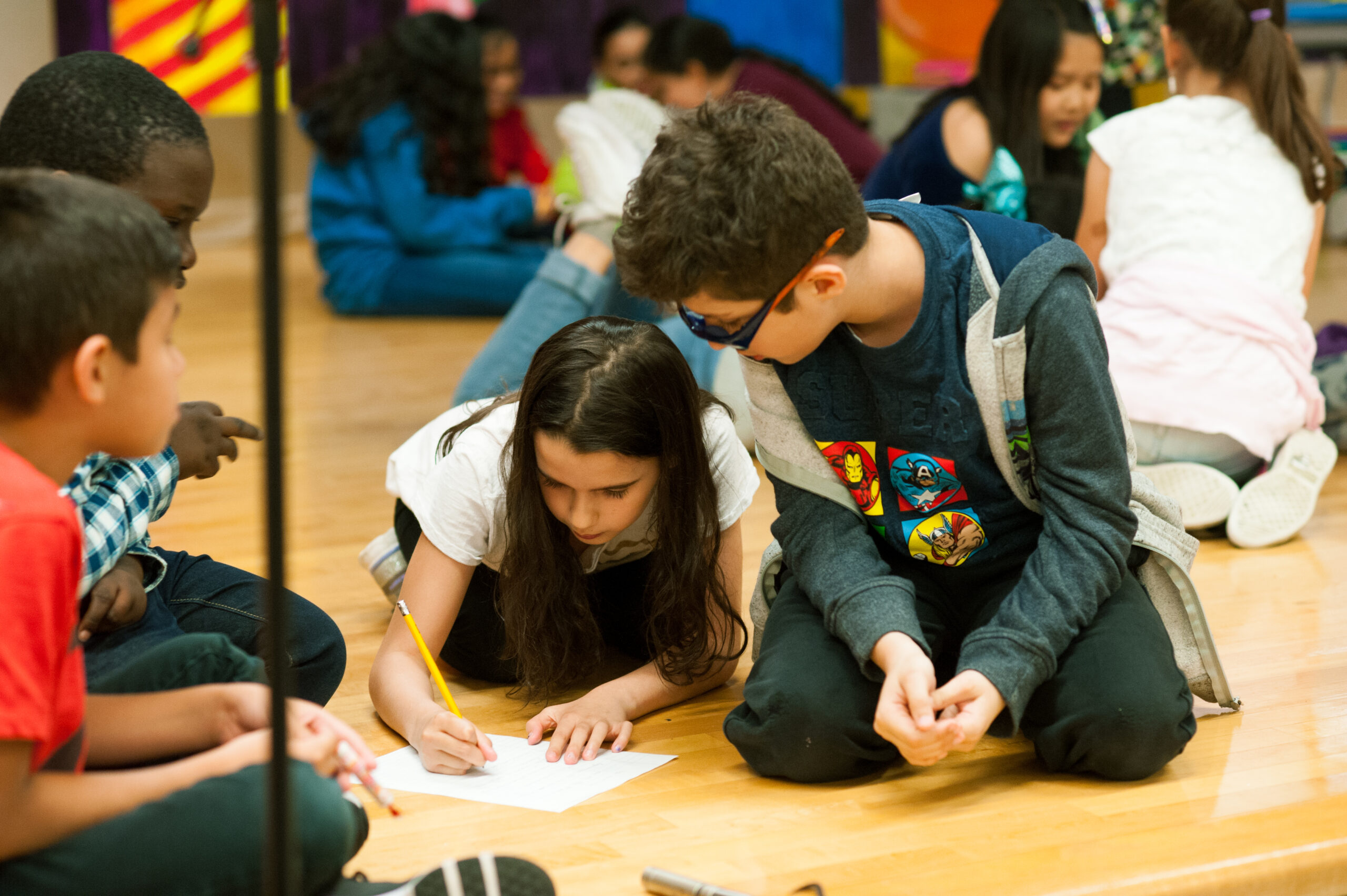 A group of 4 elementary school students sit on the floor working on a script. The student in the middle has a paper and pencil that she is using to write, as her collaborators look on.