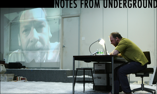 A projection screen shines a big screen on the wall of the man at a desk looking into a computer screen. With the words Notes from Underground across the top right.