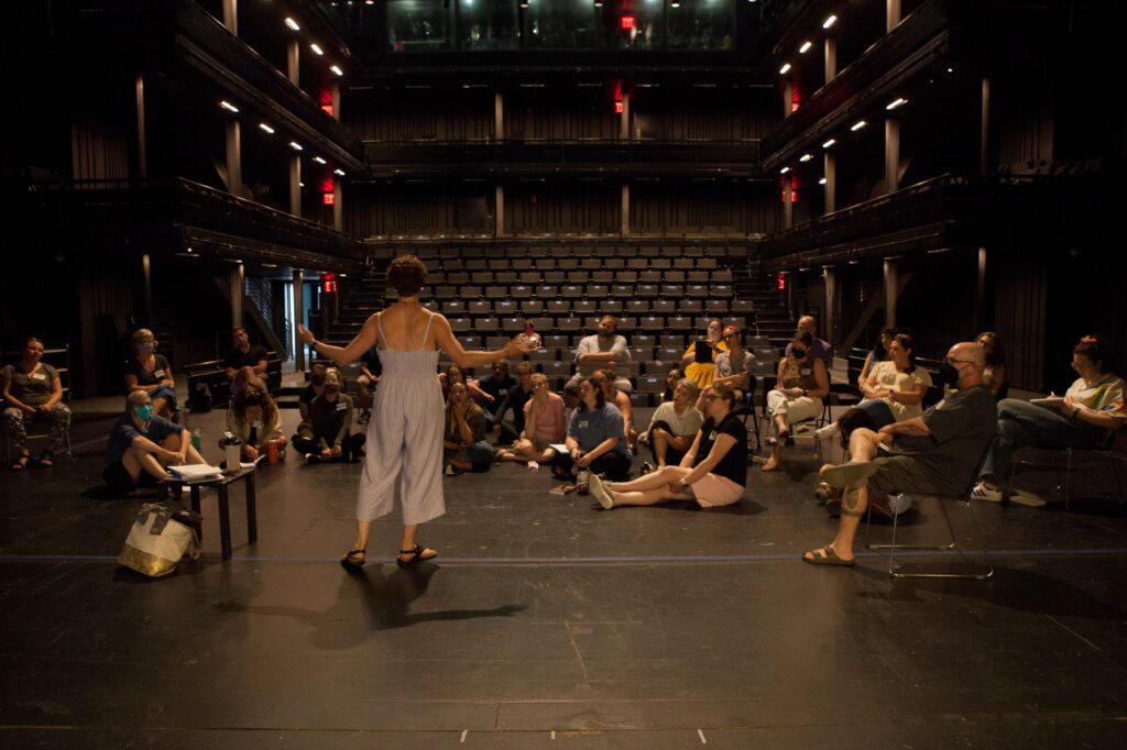 Performance faculty leads participants in a session inside the theatre at the Polonsky Shakespeare Center. She stands onstage with arms stretched, surrounded by participants seated onstage, with a view of the theatre seats behind them.