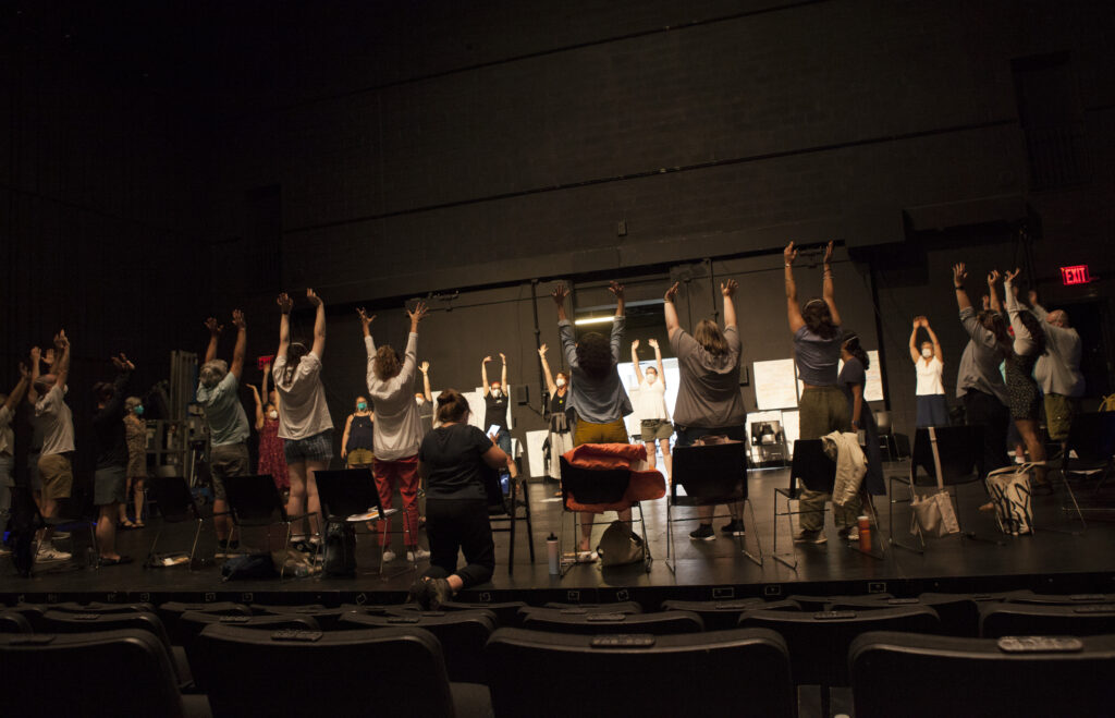 Participants stand in a circle onstage with arms raised, engaging in a warm-up activity led by performance faculty.