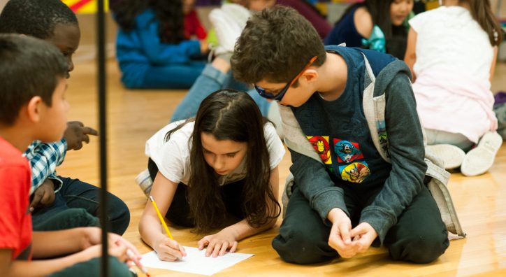 A group of 4 elementary school students sit on the floor working on a script. The student in the middle has a paper and pencil that she is using to write, as her collaborators look on.